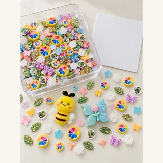Illustrative craft tray with bee, rainbow and flower pieces beside a blank card and loose focal charms